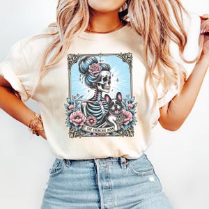 May include: A cream-colored t-shirt featuring a graphic print of a skeleton with a high bun and a pink rose, holding a French Bulldog. The design is framed in an ornate border with blue sunburst rays and pink flowers, with the text THE FRENCHIE MOM.