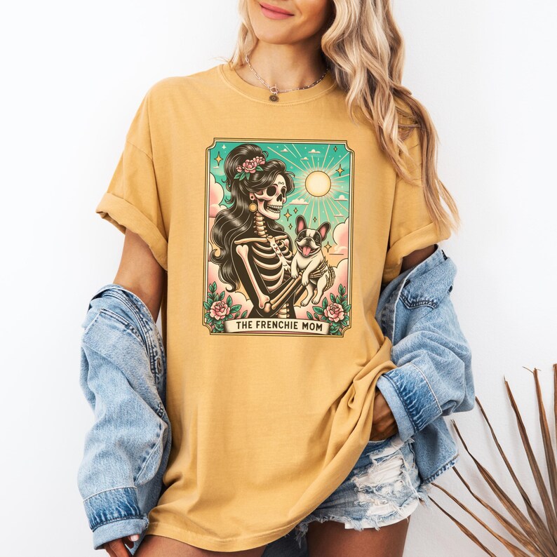 Pu&ograve; includere: Una maglietta gialla con una grafica di una donna scheletro che tiene un bulldog francese. La grafica &egrave; in stile vintage con un bordo floreale e il testo "The Frenchie Mom".