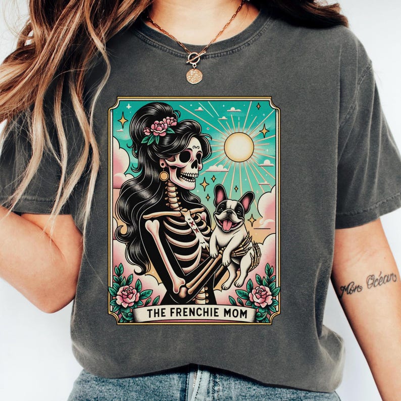 Pu&ograve; includere: Una maglietta grigio scuro con un'illustrazione in stile vintage di una donna scheletro che tiene un bulldog francese. L'illustrazione &egrave; circondata da un bordo turchese e il testo "THE FRENCHIE MOM" &egrave; stampato sotto.