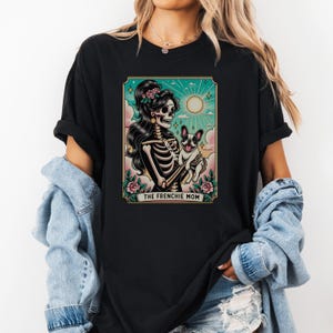 Pu&ograve; includere: Maglietta nera con una grafica di una donna scheletro che tiene un bulldog francese. La grafica &egrave; in stile vintage e ha il testo "The Frenchie Mom" in basso.