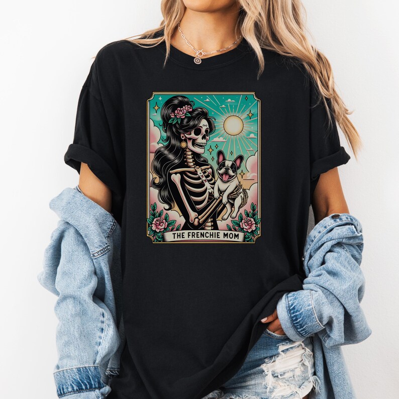 Pu&ograve; includere: Maglietta nera con una grafica di una donna scheletro che tiene un bulldog francese. La grafica &egrave; in stile vintage e ha il testo "The Frenchie Mom" in basso.