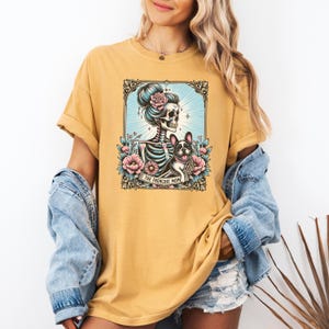 May include: A mustard yellow t-shirt featuring a graphic print of a skeleton with a hair bun and a French Bulldog surrounded by pink flowers and blue sunburst rays. A banner at the bottom of the illustration contains the text THE FRENCHIE MOM.