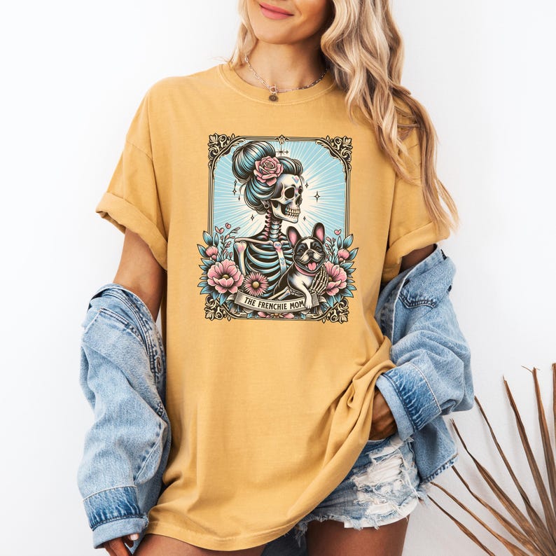 May include: A mustard yellow t-shirt featuring a graphic print of a skeleton with a hair bun and a French Bulldog surrounded by pink flowers and blue sunburst rays. A banner at the bottom of the illustration contains the text THE FRENCHIE MOM.