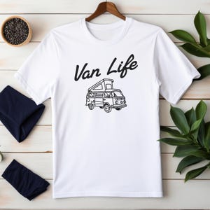 May include: White t-shirt with a black graphic of a camper van and the words "Van Life" in a script font. The shirt is hanging on a wooden hanger.
