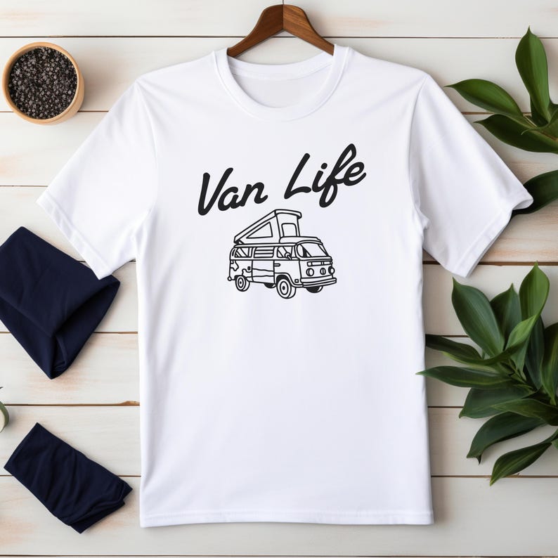 May include: White t-shirt with a black graphic of a camper van and the words "Van Life" in a script font. The shirt is hanging on a wooden hanger.