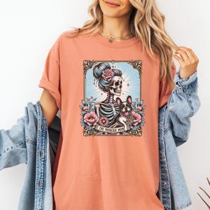 May include: A coral-colored t-shirt featuring a graphic print of a skeleton with a high bun hairstyle adorned with a pink rose, standing next to a French Bulldog. The design includes floral accents, a decorative frame, and the text THE FRENCHIE MOM.