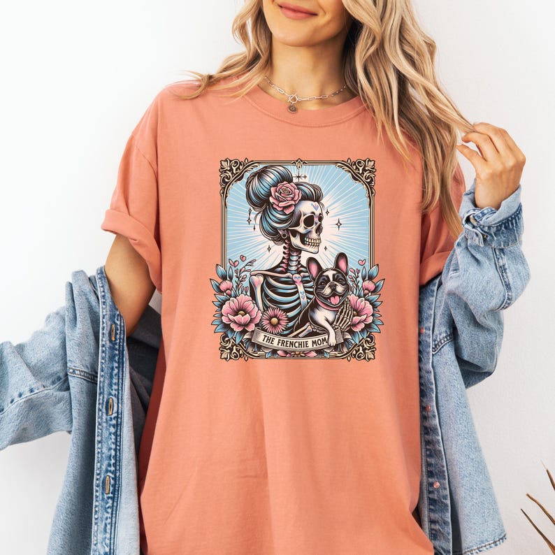 May include: A coral-colored t-shirt featuring a graphic print of a skeleton with a high bun hairstyle adorned with a pink rose, standing next to a French Bulldog. The design includes floral accents, a decorative frame, and the text THE FRENCHIE MOM.