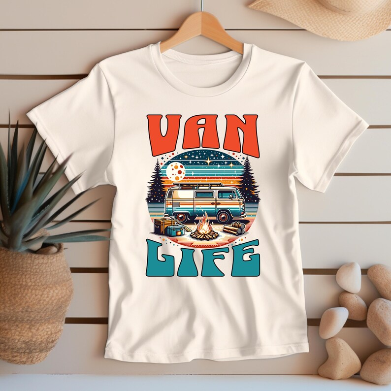 May include: White t-shirt with a retro graphic featuring a blue and orange van, campfire, and the words "VAN LIFE" in orange and teal. The design evokes a vintage camping theme.