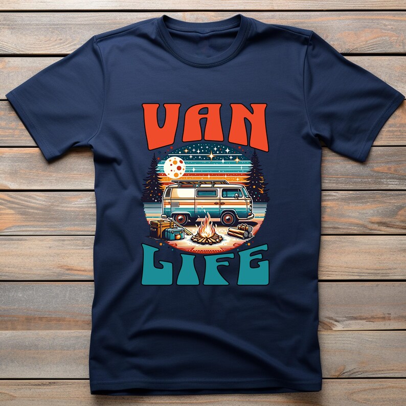 May include: Navy blue t-shirt with a colorful graphic design. The design features a retro camper van, a campfire, and the words "VAN LIFE" in orange and teal. The background includes a sunset and trees.