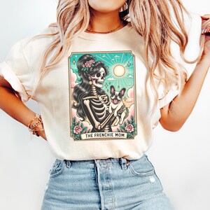 Pu&ograve; includere: Una maglietta bianca con una grafica di una donna scheletro che tiene un bulldog francese. La grafica &egrave; in stile vintage con uno sfondo colorato e il testo "The Frenchie Mom".