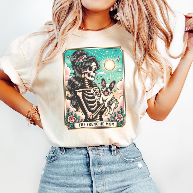 Pu&ograve; includere: Una maglietta bianca con una grafica di una donna scheletro che tiene un bulldog francese. La grafica &egrave; in stile vintage con uno sfondo colorato e il testo "The Frenchie Mom".