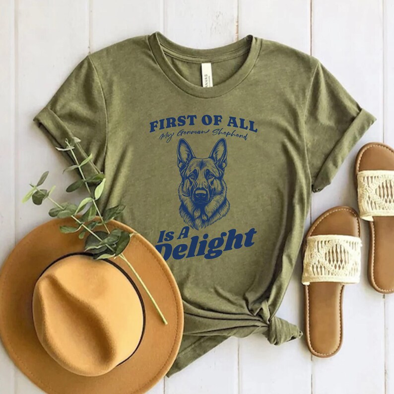 Pu&ograve; includere: Una maglietta verde con il testo "FIRST OF ALL My German Shepherd Is A Delight" in lettere blu. Un pastore tedesco &egrave; raffigurato al centro della maglietta.