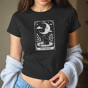 May include: Black t-shirt with a white tarot card design. The graphic shows a crescent moon with a face, a black cat, and the words "THE MOON". The design has a detailed border and botanical elements.