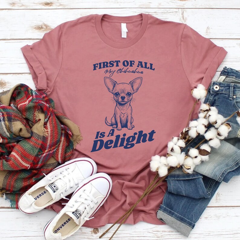 May include: A pink t-shirt with the text "First of all, my chihuahua is a delight" in blue lettering. A chihuahua illustration is centered on the shirt.