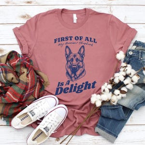 Pu&ograve; includere: Una maglietta rosa con il testo "FIRST OF ALL My German Shepherd Is A Delight" in lettere blu. Un'illustrazione di un pastore tedesco &egrave; presente sulla maglietta.