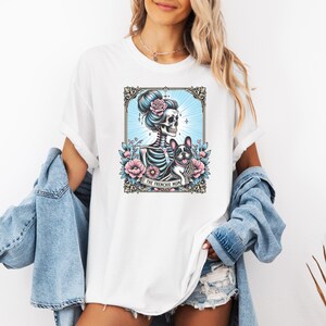 May include: A white t-shirt featuring a graphic print of a skeleton with a high bun and a French bulldog surrounded by pink flowers and blue rays within an ornate frame. A banner at the bottom of the graphic contains the text THE FRENCHIE MOM.