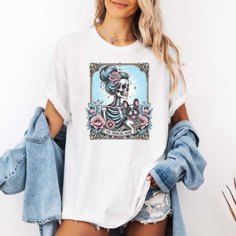 May include: A white t-shirt featuring a graphic print of a skeleton with a high bun and a French bulldog surrounded by pink flowers and blue rays within an ornate frame. A banner at the bottom of the graphic contains the text THE FRENCHIE MOM.