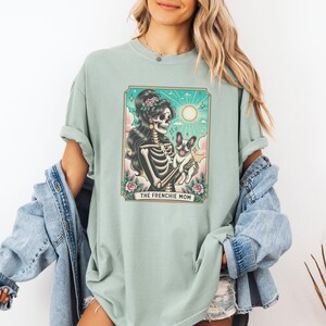 Pu&ograve; includere: Una maglietta verde chiaro con una stampa grafica di una donna scheletro che tiene un bulldog francese. La grafica &egrave; in stile vintage e include il testo "The Frenchie Mom".