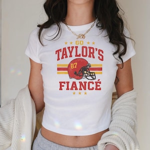 May include: White t-shirt with a vintage-style graphic. The design features the text "GO TAYLOR'S FIANCÉ" with a football helmet illustration. The helmet is red and gold, with the number 87.