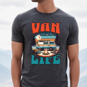 May include: Dark gray t-shirt with a colorful graphic. The graphic features a vintage van, campfire, and the words "VAN LIFE" in orange and teal. The design evokes a camping and travel theme.