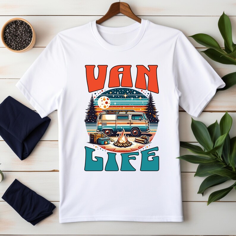 May include: White t-shirt with a retro graphic design. The design features a blue and orange van, a campfire, and the words "VAN LIFE" in orange and teal. The shirt is displayed on a wooden hanger.