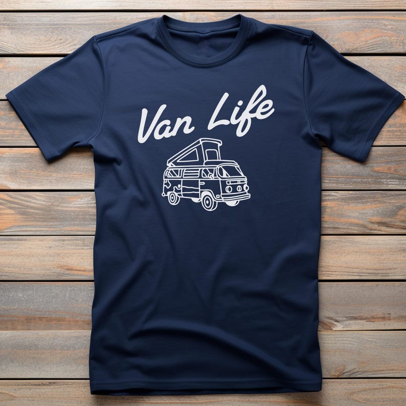 May include: Navy blue t-shirt with the white text "Van Life" above a white line drawing of a camper van. The shirt has short sleeves and a crew neck. The background is a wooden surface.