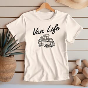 May include: Cream-colored t-shirt featuring a black graphic of a camper van with the text "Van Life" above it. The van has an open top and is detailed with windows and wheels. The shirt is hanging on a wooden hanger.