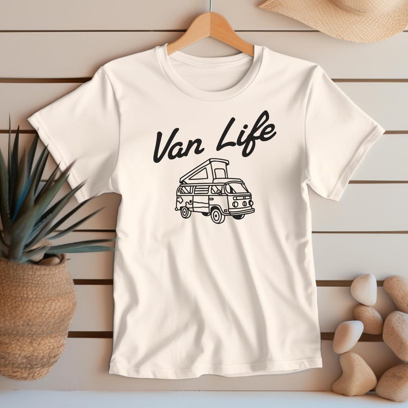 May include: Cream-colored t-shirt featuring a black graphic of a camper van with the text "Van Life" above it. The van has an open top and is detailed with windows and wheels. The shirt is hanging on a wooden hanger.