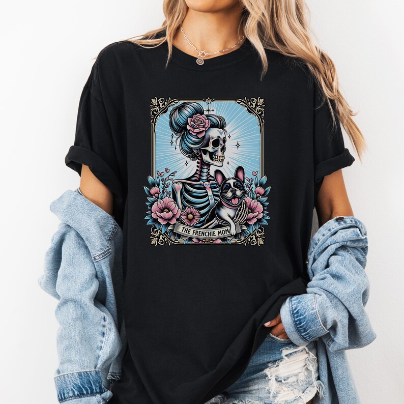 May include: A black t-shirt featuring a graphic illustration of a skeleton with a pink rose in its hair, standing next to a French Bulldog. The design is framed by flowers and includes a banner at the bottom with the text, THE FRENCHIE MOM.