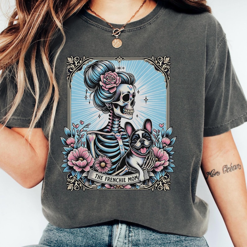 May include: A charcoal gray t-shirt featuring a graphic print of a skeleton with a pink rose in its hair, holding a French bulldog. The design is framed in an ornate border with pink and blue flowers and the text THE FRENCHIE MOM at the bottom.