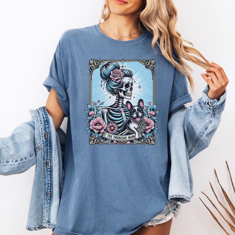 May include: A blue t-shirt featuring a graphic print of a skeleton with a hair bun and a French Bulldog. The design includes pink flowers, a light blue sunburst background, and a banner at the bottom with the text THE FRENCHIE MOM.