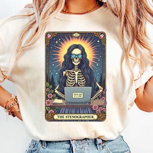 Peut inclure: Un t-shirt blanc avec un graphique d'un squelette portant des lunettes de soleil et tapant sur un ordinateur portable. Le graphique est dans le style d'une carte de tarot et dit "The Stenographer" et "Speak One At A Time".