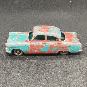 Könnte beinhalten: Ein Vintage-Spielzeugauto aus Metall, lackiert in einem verblassten Teal- und Rot-Farbschema. Das Auto hat ein leicht verbeultes Dach und weist Gebrauchsspuren auf.