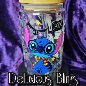 Peut inclure: Un gobelet en verre transparent avec un couvercle et une paille en bambou, orné d'un personnage de dessin animé bleu avec des oreilles roses et des lunettes. Le gobelet est décoré d'étoiles et d'autres graphiques. Le texte "Deluxious Bling" est affiché sur la base.