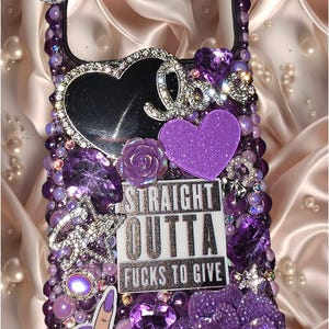 May include: A phone case decorated with purple beads, rhinestones, and charms. It features a silver bow, a black heart, a purple rose, and a rectangular plaque with the text STRAIGHT OUTTA FUCKS TO GIVE. A hand gesture decal and a large purple flower.