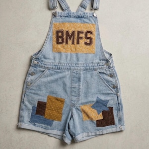 May include: Light blue denim overall shorts with patches and the letters "BMFS" on the bib. The patches are various colors and patterns, including a star shape. The shorts have adjustable straps and button closures.