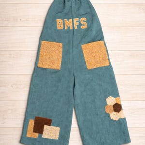 May include: Teal corduroy overalls with the letters "BMFS" in gold. The overalls have two large square pockets and decorative patches in shades of brown and tan. A floral patch is on the right leg.