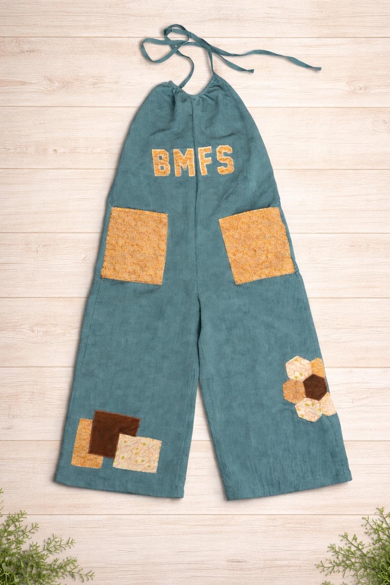 Puede incluir: Mono de pana color turquesa con las letras "BMFS" en dorado. El mono tiene dos bolsillos cuadrados grandes y parches decorativos en tonos marrones y beige. Un parche floral est&aacute; en la pierna derecha.