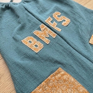 Puede incluir: Mono de color turquesa con las letras "BMS" aplicadas en tela dorada texturizada. El mono tiene una tela texturizada y un bolsillo con estampado floral dorado.