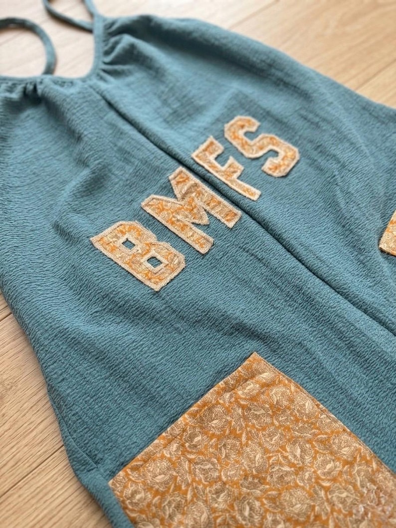 Puede incluir: Mono de color turquesa con las letras "BMS" aplicadas en tela dorada texturizada. El mono tiene una tela texturizada y un bolsillo con estampado floral dorado.