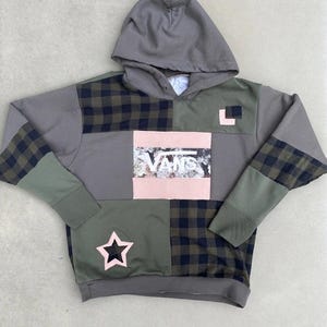Op de afbeelding: Een hoodie met een patchworkontwerp in olijfgroen, antracietgrijs en een donkergroen met zwart ruitpatroon. Het midden toont een rechthoekige patch met de witte tekst VANS over een bloemenprint, geaccentueerd door een roze ster.