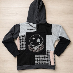 Könnte beinhalten: Ein Patchwork-Hoodie in Schwarz, Grau und Rosa. Auf der Vorderseite befindet sich eine Totenkopf-Grafik auf einem schwarzen Quadrat. Die Ärmel sind schwarz und grau, die Kapuze dunkelgrau. Das Design integriert verschiedene Texturen und Muster.