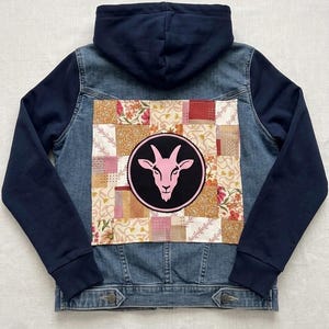 May include: A denim jacket with a navy blue hood and sleeves. The back features a patchwork design with floral and geometric patterns, and a pink goat head emblem in a black circle.
