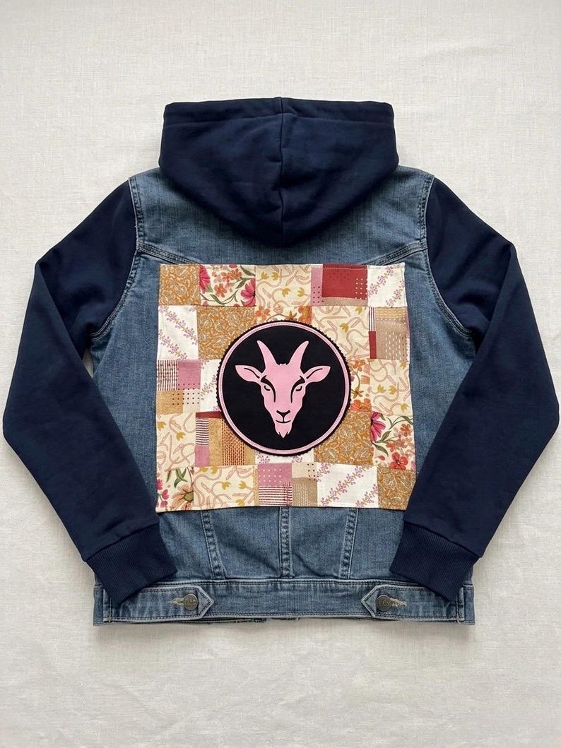 Pode incluir: Casaco de ganga com capuz e mangas azul marinho. As costas apresentam um design de patchwork com padr&otilde;es florais e geom&eacute;tricos, e um emblema de cabe&ccedil;a de cabra rosa num c&iacute;rculo preto.