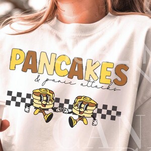 Könnte beinhalten: Weißes T-Shirt mit einem Aufdruck von zwei Cartoon-Pfannkuchen mit Gesichtern und dem Text "PANCAKES & panic attacks" in braunen und gelben Buchstaben.