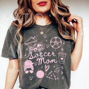 May include: A gray t-shirt with a pink graphic design that says "Soccer Mom" with soccer-related illustrations like a soccer ball, a car, a whistle, and a coffee cup.