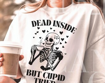 May include: White t-shirt with a black skeleton graphic holding a cup of coffee. The text "Dead Inside But Cupid Tried" is printed on the shirt.