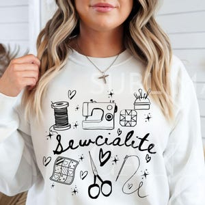 Puede incluir: Sudadera blanca con un diseño gráfico negro que incluye una máquina de coser, tijeras, hilo y el texto "Sewcialite".