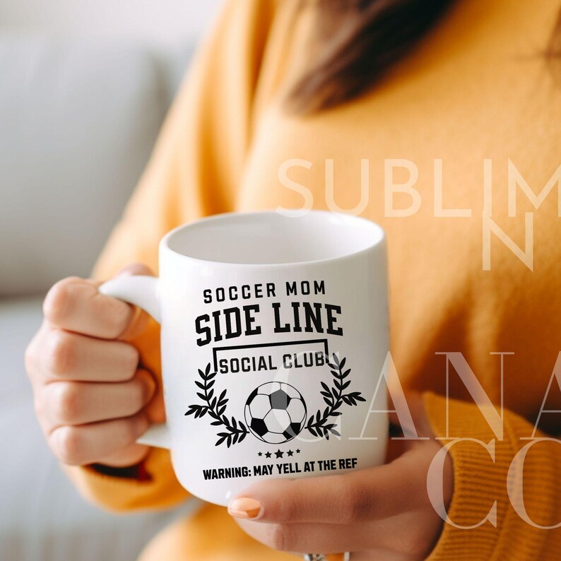 Puede incluir: Taza de cer&aacute;mica blanca con un dise&ntilde;o negro que dice "Soccer Mom Side Line Social Club" con un bal&oacute;n de f&uacute;tbol y una corona de laurel. El texto debajo del dise&ntilde;o dice "Advertencia: Puede gritar al &aacute;rbitro."