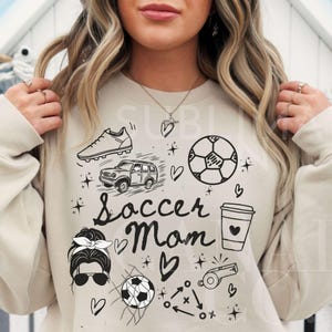 May include: A beige sweatshirt with a black graphic design that says "Soccer Mom" and includes various soccer-related images, including a soccer ball, a soccer cleat, a car, a coffee cup, a whistle, and a soccer net.